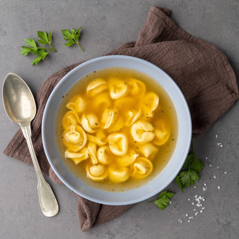 A light blue bowl of tortellini in broth, garnished with parsley, on a gray surface.