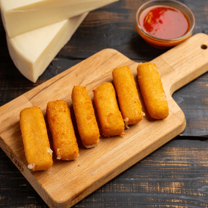 Six golden-brown mozzarella sticks on a wooden board, with a bowl of marinara sauce and cheese in the background.